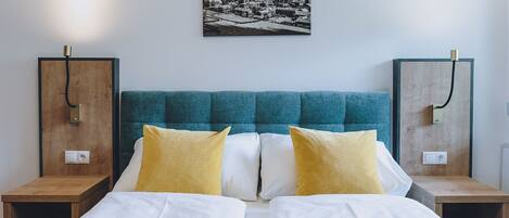 A double bed with white bedding, two yellow pillows, and a teal upholstered headboard. There are two wooden nightstands with brass fixtures on either side of the bed. A black and white photograph of a snowy mountain landscape hangs on the wall above the bed.