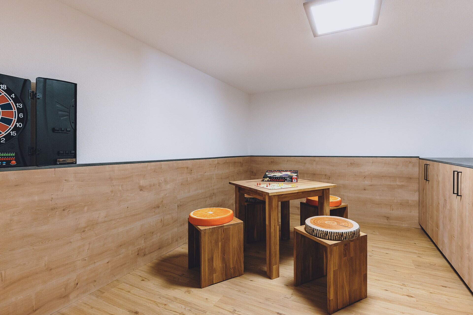 Una habitación de esquina con piso de madera, una pequeña mesa con un juego de mesa, dos taburetes y un reloj montado en la pared.