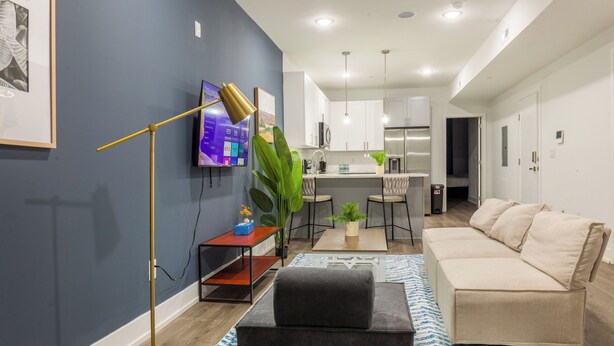 Una sala moderna con un sofá, una mesa de centro y un televisor. Se observa una cocina al fondo con armarios blancos y un comedor.