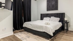 Un dormitorio moderno con cabecera negra, ropa de cama blanca, televisor de pantalla plana montado en la pared y una alfombra estampada en el suelo.