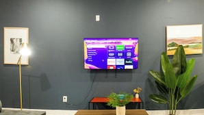 Una sala de estar con un televisor de pared mostrando la interfaz de un servicio de streaming, una planta en maceta y un cuadro en la pared.
