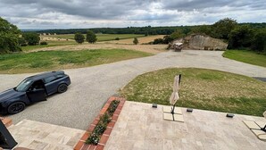 A driveway leading to a house with a car parked on the left. There is a patio area with a table and chairs, and a brick planter with flowers. - Le Clos de Galou, Authenticity, Comfort, Nature (Aucamville)