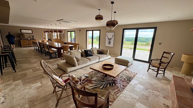 A spacious living area with a large sofa, dining table, and chairs, a kitchen in the background, and large windows offering a view of the outdoors. - Le Clos de Galou, Authenticity, Comfort, Nature (Aucamville)