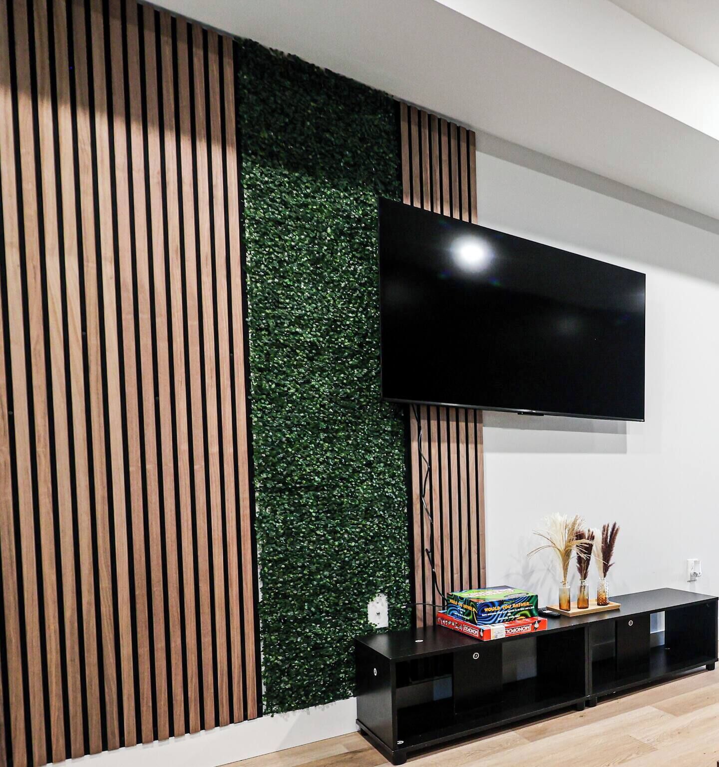 A modern living room with a flat-screen TV mounted on the wall, a black entertainment unit below, and a vertical wooden panel on the left.