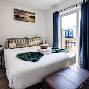 A hotel room with a bed, a bench, a window with curtains, a wall-mounted painting, and a view of buildings outside. - Sun-filled 2BR/1BA close to the Philadelphia Zoo and UPenn. Enjoy central air, in-unit laundry, and a sleek, modern kitchen — comfortably sleeps 6. Easy access to I-76 and I-676 for getting around the city. (Philadelphia)