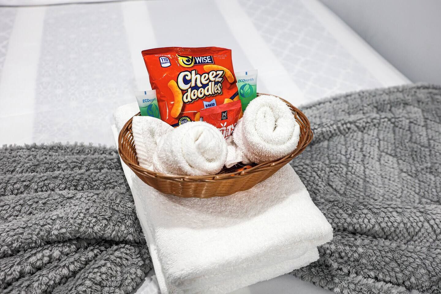 A basket with white towels and a pack of Wise Cheez doodles on a white towel on a bed.