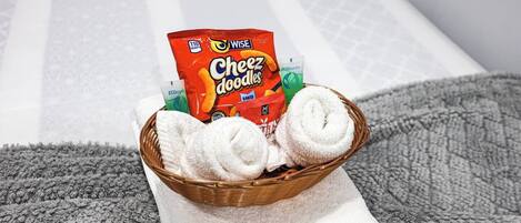 A basket with white towels and a pack of Wise Cheez doodles on a white towel on a bed.