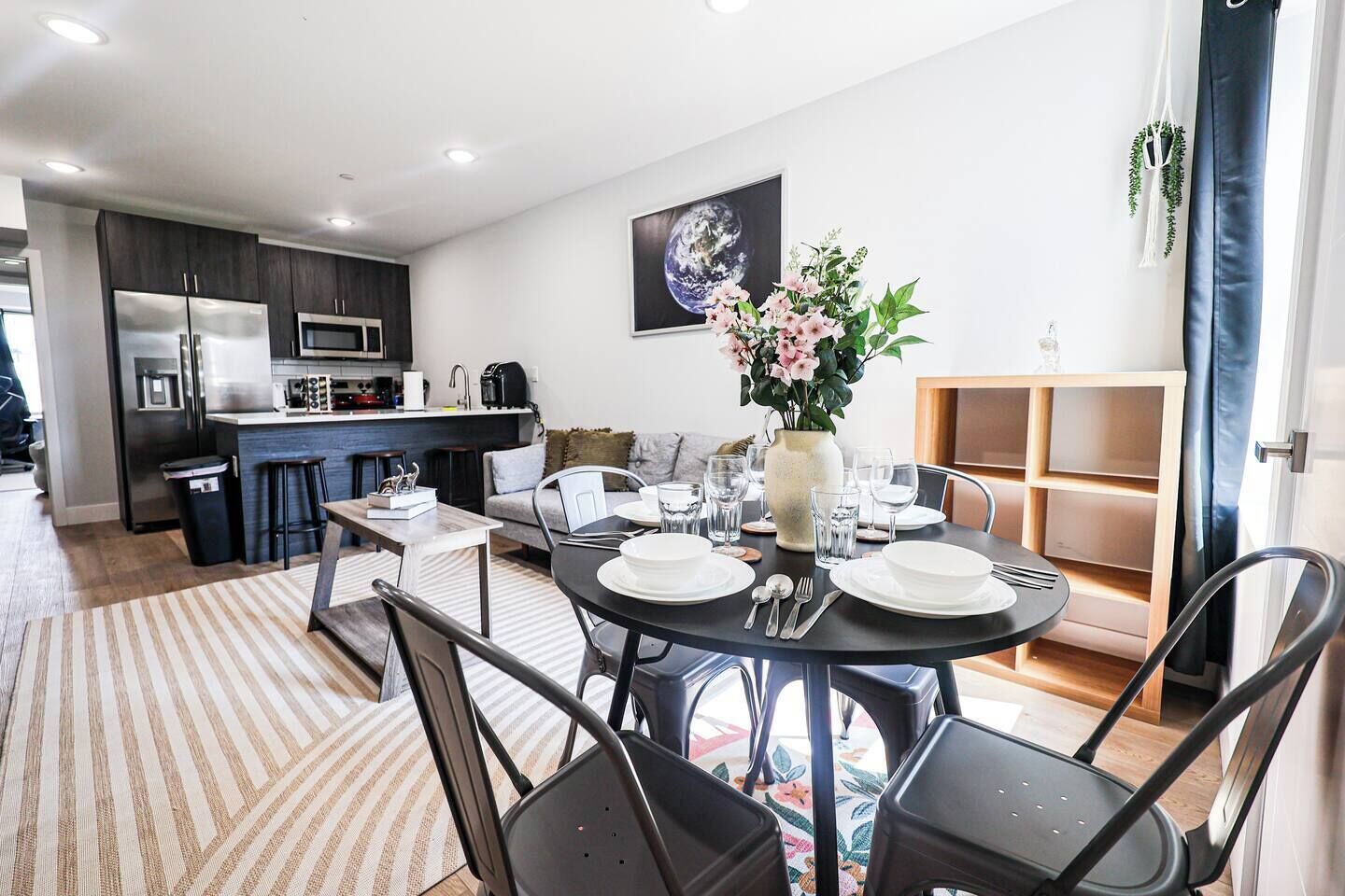 A modern kitchen with dark cabinets and stainless steel appliances, a dining area with a round table set for four, and a living space with a sofa and a large painting of Earth.