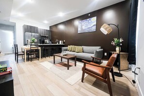 A modern living room with a grey sofa, wooden coffee table, and a brown leather chair. A kitchen with dark cabinetry and a dining area with a wooden table and chairs are visible. - Modern 2BR near Philly Zoo & UPenn! Features central air, in-unit laundry, sleek kitchen & sleeps 6. Quick access to I-76/676 & top attractions. (Philadelphia)