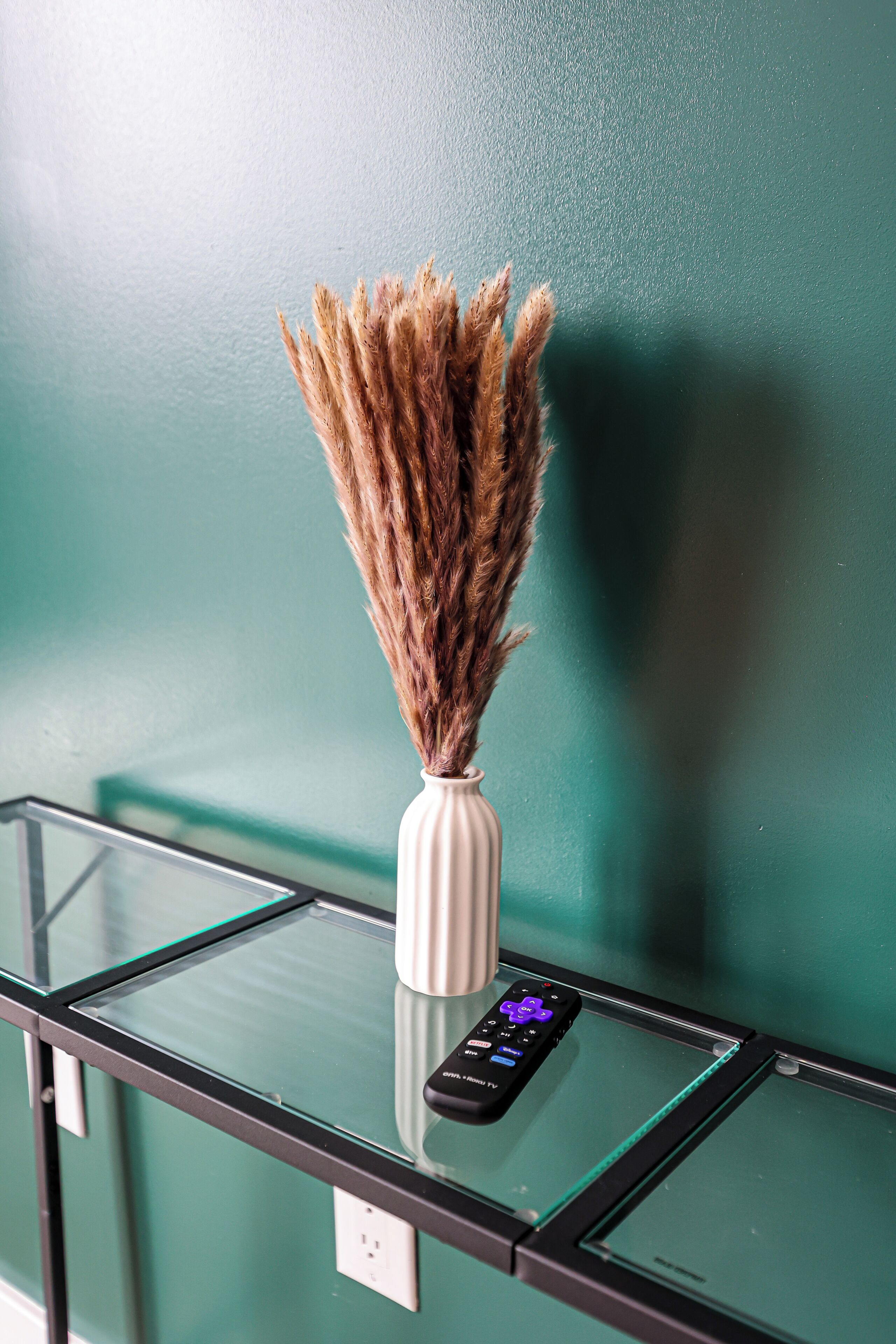 A glass table with a white vase containing dried pampas grass, a remote control, and a power outlet.