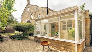 A conservatory with stone walls and large windows, a gravel path, and a wooden stool. - Beautiful 2BD Cottage in the Cotswolds (Bourton-on-the-Hill)