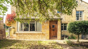 A stone house with a wooden door, a window, and a small garden area with a tree and a hedge. - Beautiful 2BD Cottage in the Cotswolds (Bourton-on-the-Hill)
