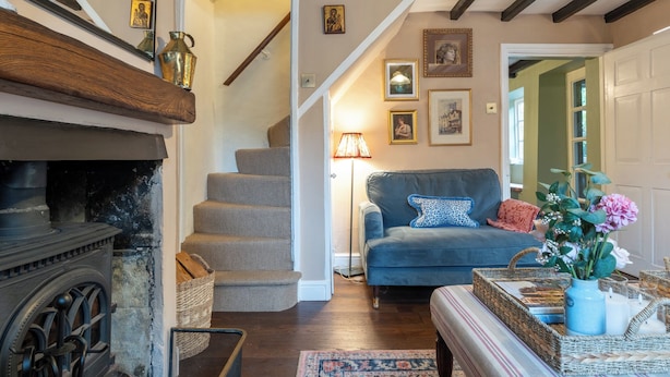 A cozy living room with a fireplace, a blue sofa, and a staircase leading to another room. - Beautiful 2BD Cottage in the Cotswolds (Bourton-on-the-Hill)