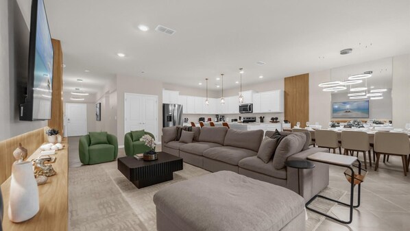 A modern living room with a large sofa, a coffee table, and a dining area with a table set for four. - Stunning Resort Villa w Pool, Cinema & Games Room (Clermont)