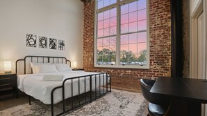 Ein Schlafzimmer mit Bett, Schreibtisch und großem Fenster mit Blick auf einen rosafarbenen Himmel.