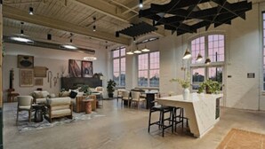 A spacious living area with a kitchen, large windows, and a high ceiling with exposed beams.