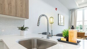 A modern kitchen with a stainless steel sink, wooden cabinets, a potted plant, a bar stool, and a bottle of lotion on the counter.
