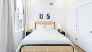 A bedroom with a bed, two bedside tables with lamps, a window with curtains, and two wall art pieces.