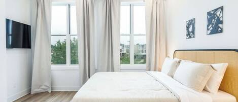A bedroom with a large bed, a flat-screen TV mounted on the wall, and a window with curtains.