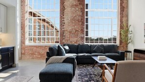 A spacious loft with exposed wooden beams, a brick wall, and large windows offering views of a building with arched windows. - Warm and Inviting Abode (Greenville)