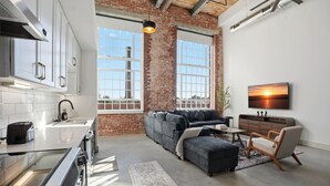 A modern kitchen with white cabinets and a stainless steel range hood, a gray sectional sofa with a throw blanket, a wooden side table, and a TV mounted on the wall. - Warm and Inviting Abode (Greenville)
