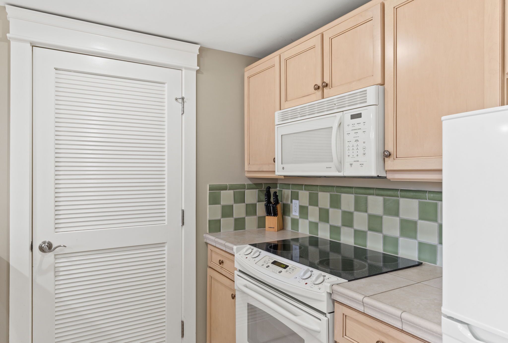 Una cocina con estufa, microondas y horno blancos, armarios de madera clara y un salpicadero de azulejos verdes y blancos.
