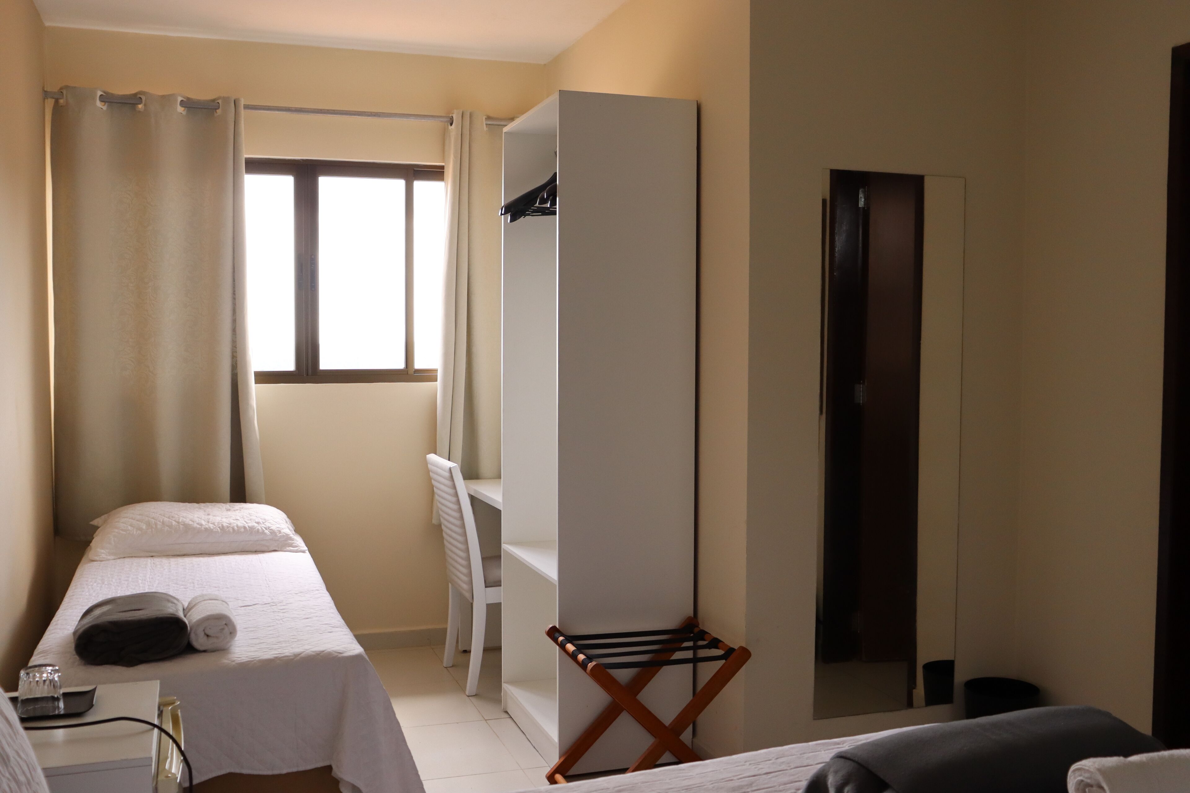 Habitación de hotel con dos camas, una ventana con cortinas, un armario, un espejo y una pequeña mesa con un vaso y una toalla.