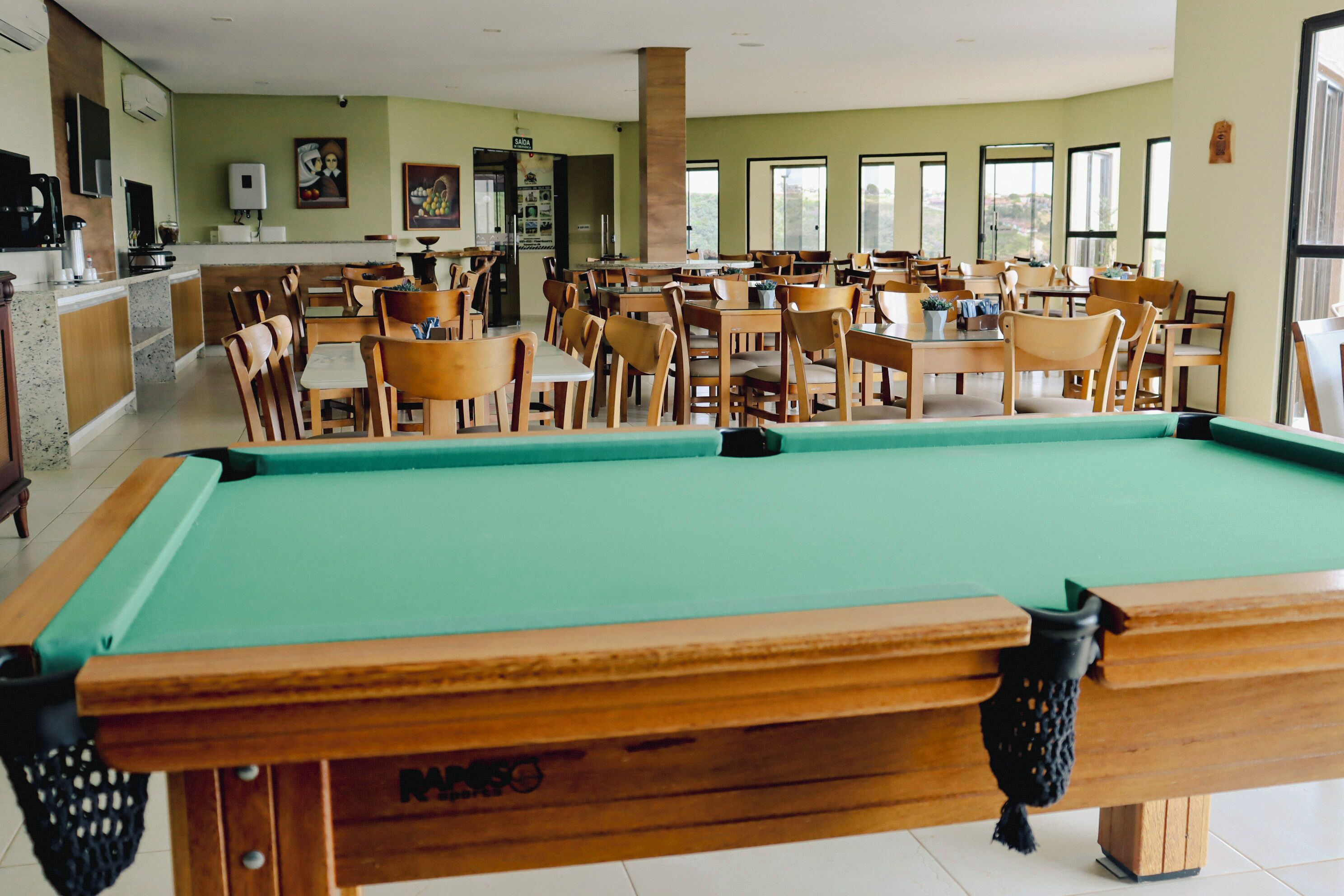 A pool table in a dining area with multiple tables and chairs, a kitchen area, and large windows.