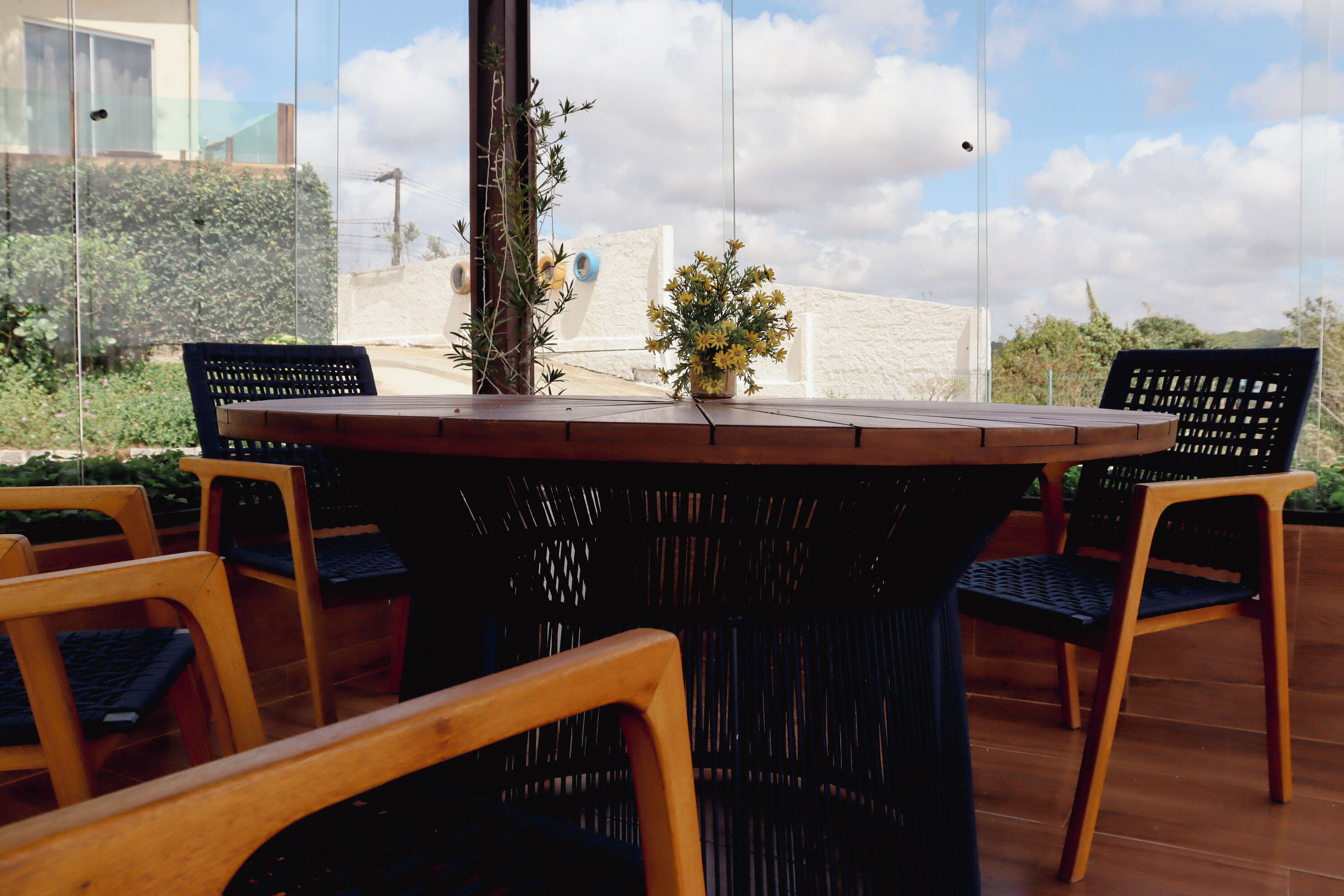A round wooden table with a vase of yellow flowers, surrounded by four wooden chairs with black cushions.