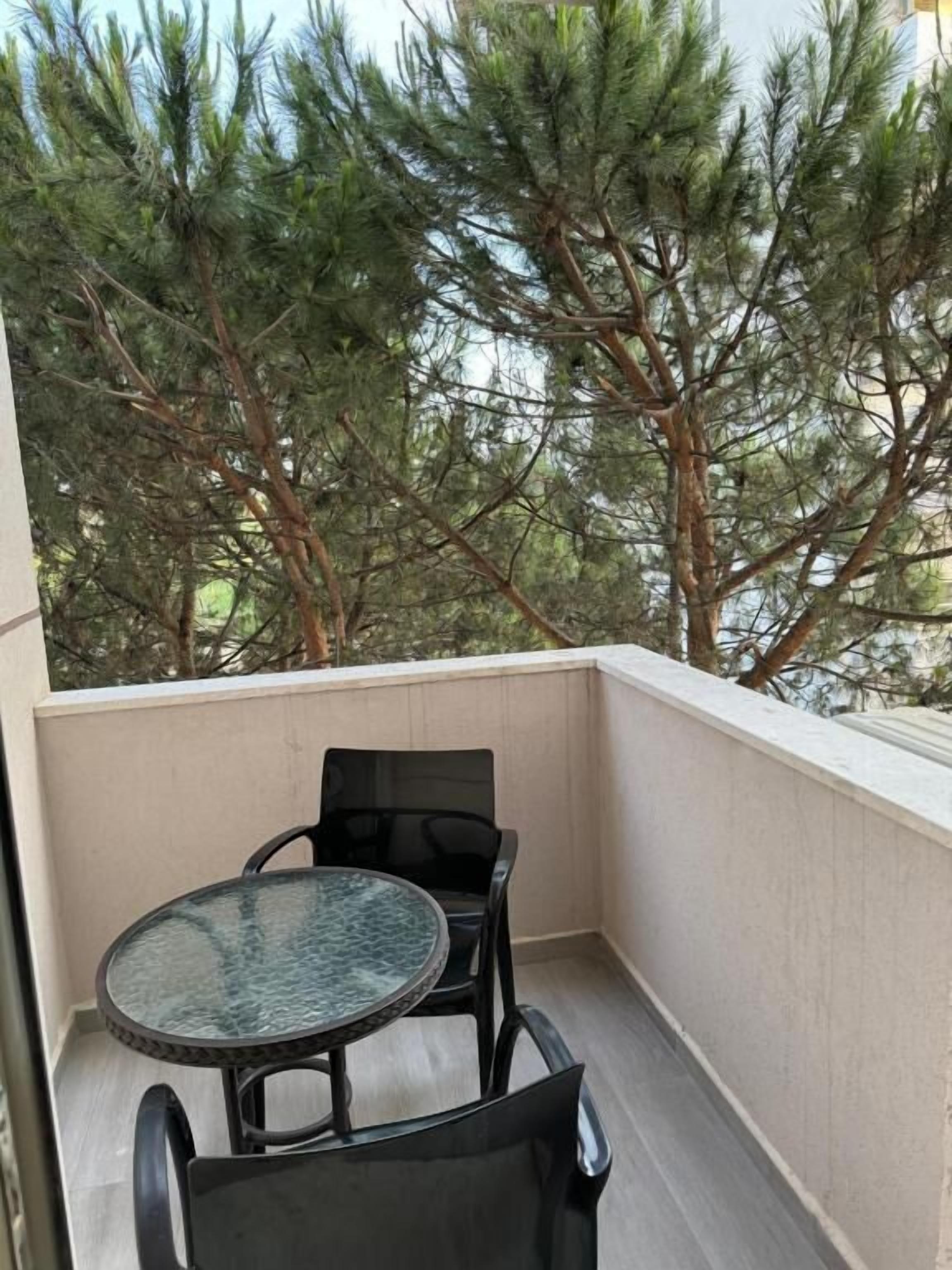 Un balcon doté d’une table en verre et de deux chaises, entouré de grands pins.