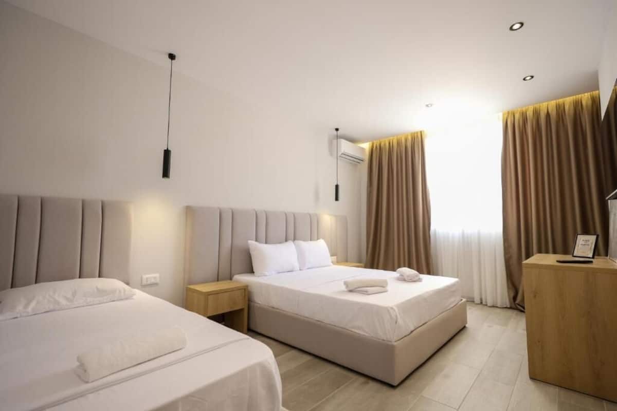Habitación de hotel con dos camas, una mesita de noche de madera, un escritorio y un ventanal con cortinas.