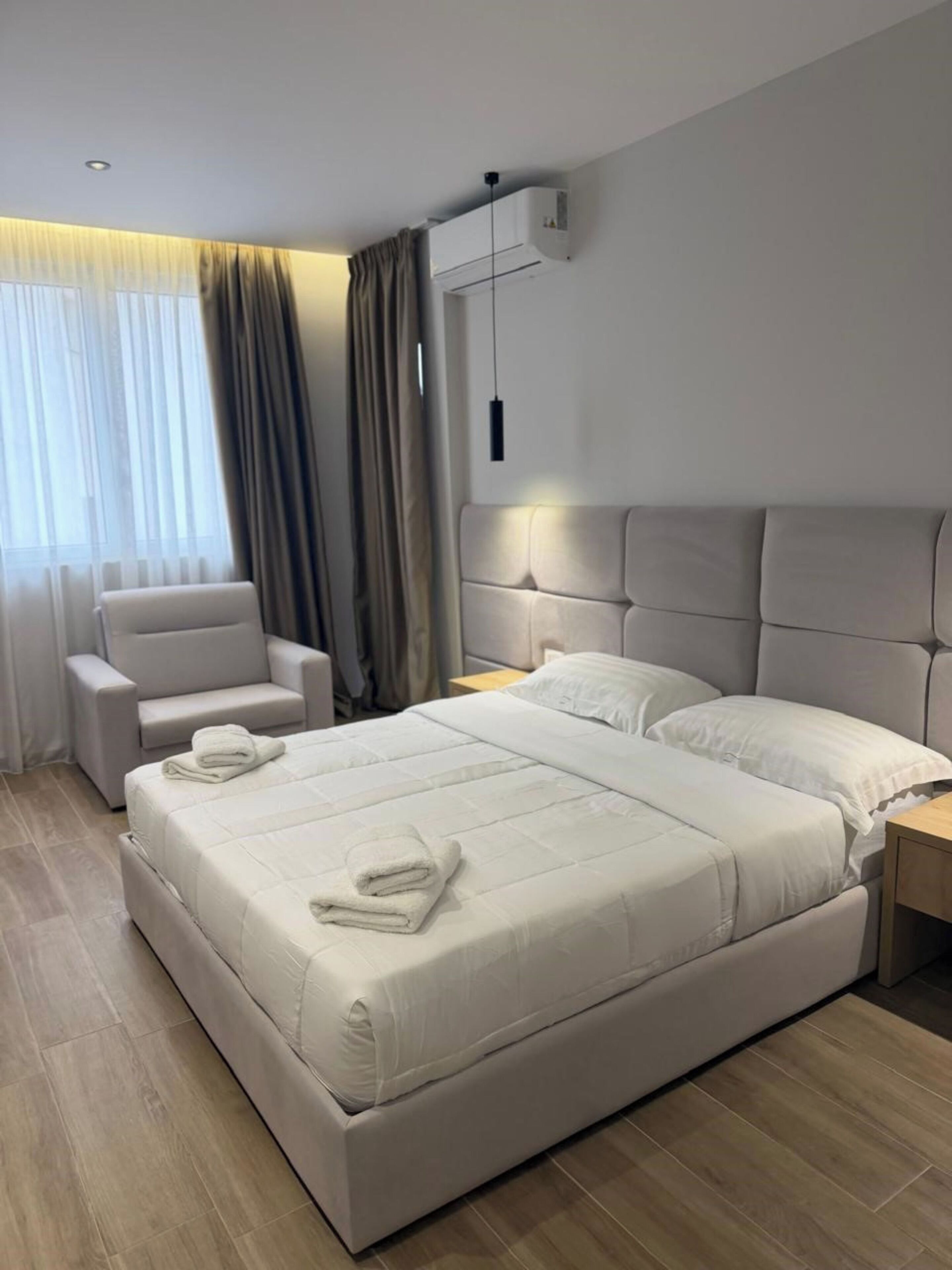 Habitació d'hotel moderna amb un llit gran, una cadira, una tauleta de nit i una finestra amb cortines.