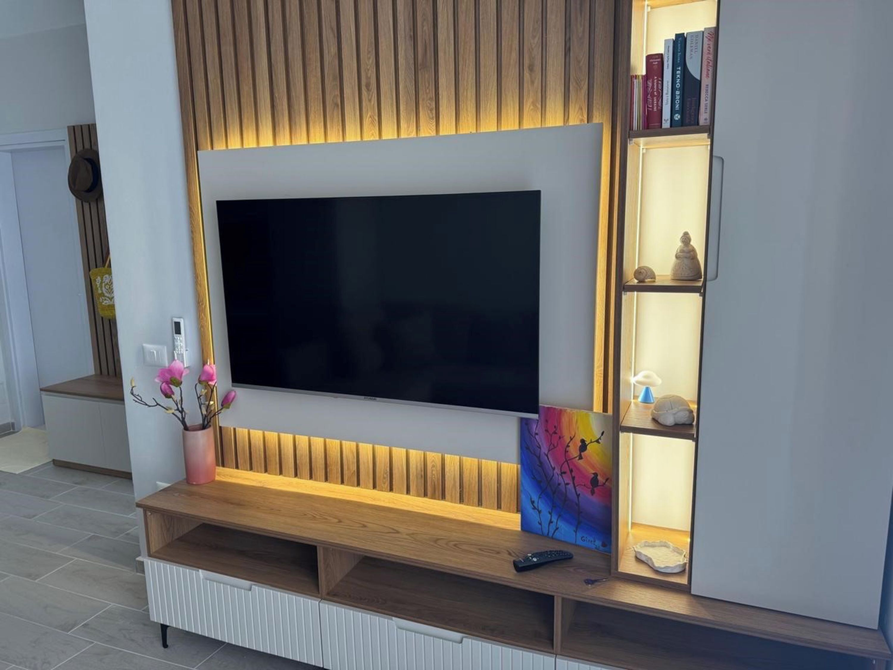 Un salon moderne doté d’un grand téléviseur à écran plat fixé à un mur en bois, d’une étagère intégrée agrémentée d’objets décoratifs et d’une petite table d’appoint.