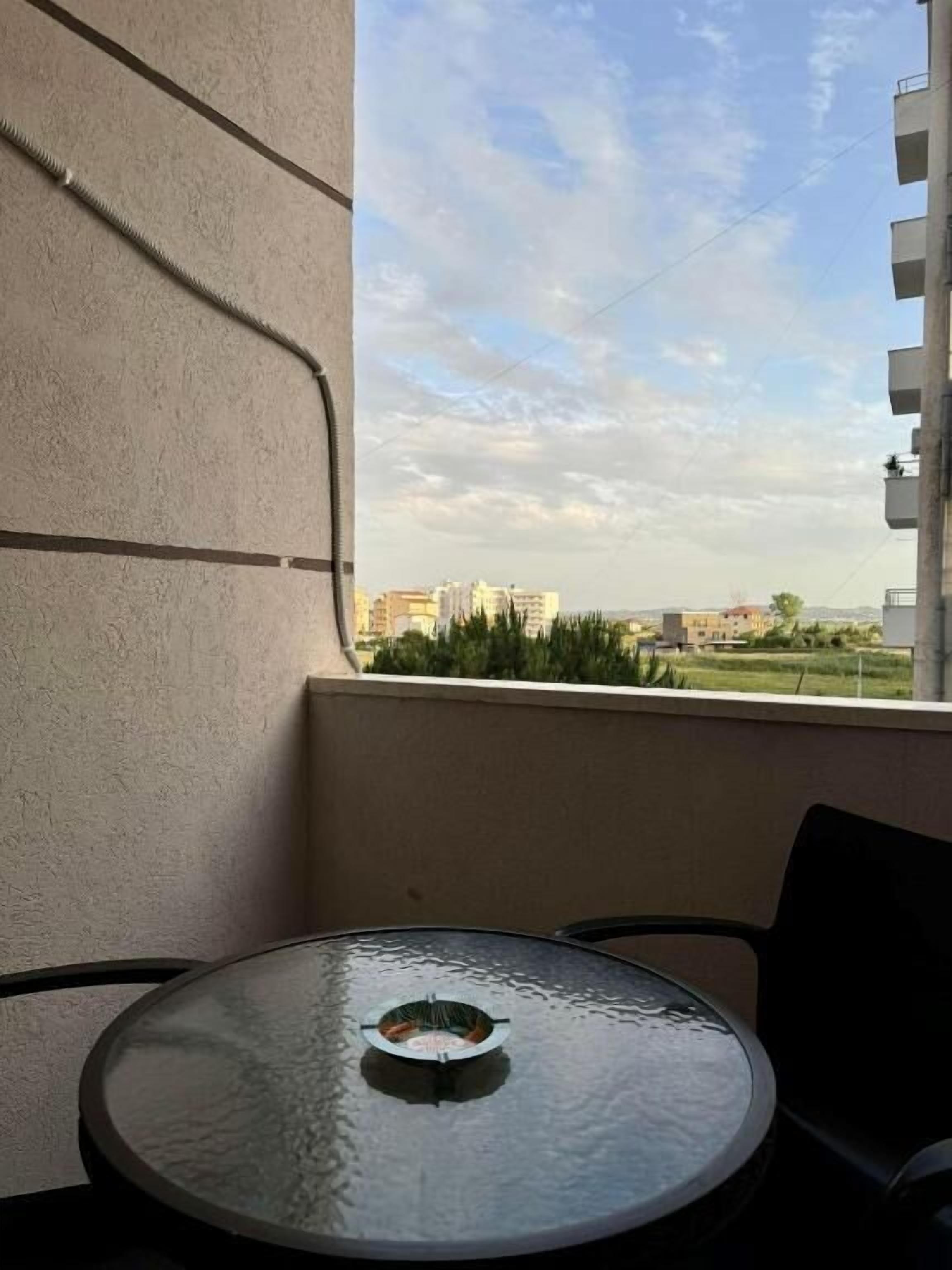 Un balcón con una mesa redonda de vidrio y sillas, con vistas a un paisaje urbano con edificios y vegetación.