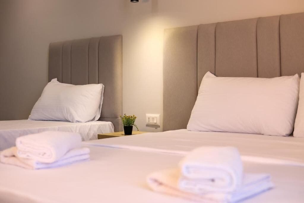 A hotel room with two beds, each with a headboard, a small bedside table with a lamp, and neatly folded towels on the beds.