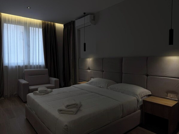 A modern hotel room with a large bed, a chair, a bedside table, and a window with curtains.