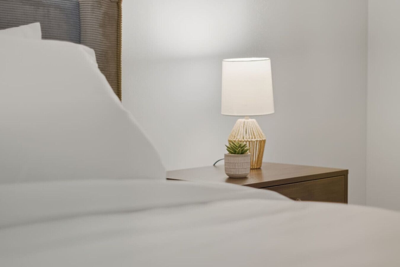 Un lit bien fait, avec du linge de lit blanc, une lampe de chevet à la base texturée et une petite plante en pot posée sur une table de chevet en bois.