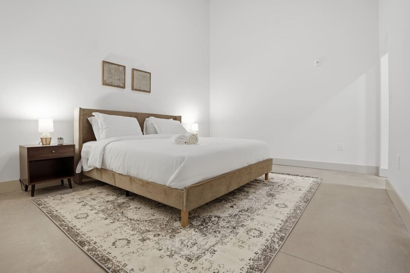 Un dormitorio con cama de madera, ropa de cama blanca, una mesita de noche con lámpara y una alfombra estampada.