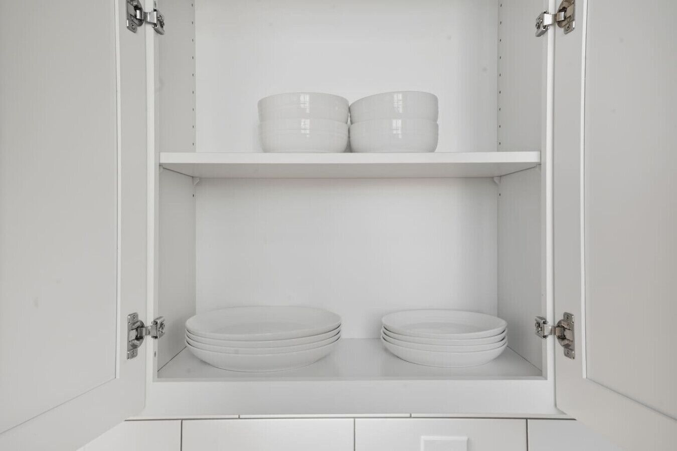Un meuble blanc à deux étagères ; l’étagère supérieure contient deux récipients blancs, et l’étagère inférieure contient plusieurs assiettes blanches.