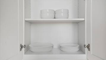 Un meuble blanc à deux étagères ; l’étagère supérieure contient deux récipients blancs, et l’étagère inférieure contient plusieurs assiettes blanches.