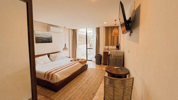 A hotel room with a bed, a small table, a chair, a TV, and a window with a view. - TWT CONDOS (Tulum)