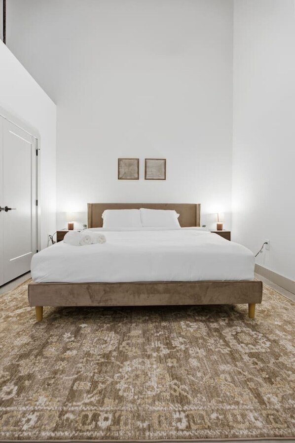 A modern bedroom with a wooden bed, white bedding, a patterned rug, and two wall-mounted lights. - Stylish + Comfortable Apt Close to DT GVL (Greenville)