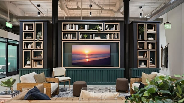 Een moderne lounge met een grote flatscreen-tv waarop een zonsondergang te zien is, omringd door boekenplanken en comfortabele zitplaatsen.