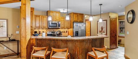 A kitchen with wooden cabinets, a central island with bar stools, a microwave, a refrigerator, and pendant lighting.