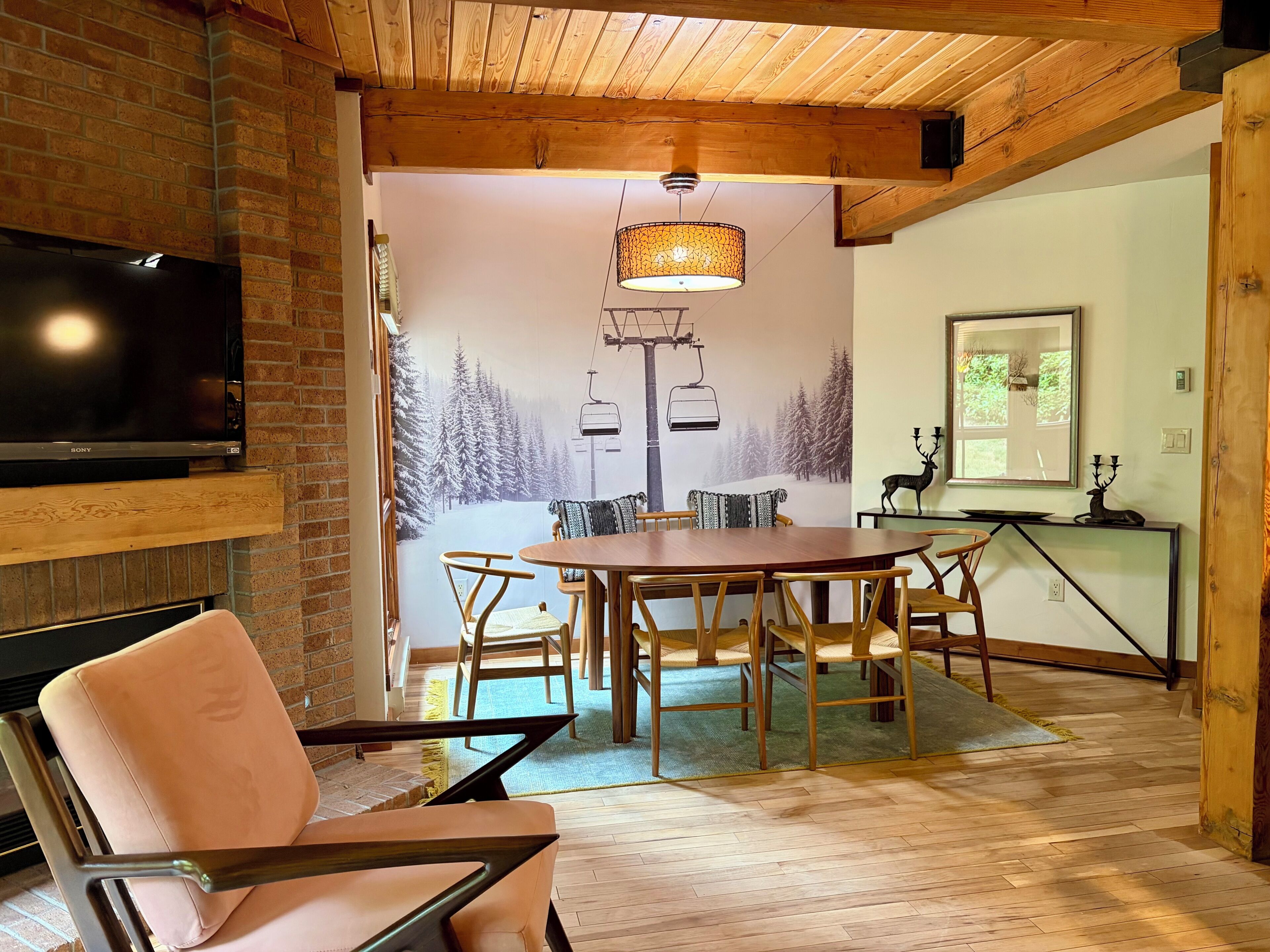 A cozy dining area with a wooden table and chairs, a television mounted on a brick wall, and a mural of a snowy mountain scene.