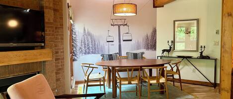 A cozy dining area with a wooden table and chairs, a television mounted on a brick wall, and a mural of a snowy mountain scene.