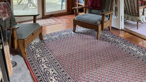Ein Holzblockhaus-Interieur mit einer großen Schiebeglastür, die auf eine Terrassensitzgelegenheit hinausführt, einem gemusterten Teppich und einem Bücherregal mit einer Lampe.
