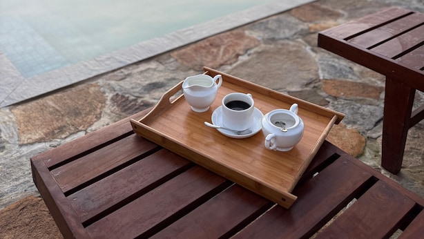 Ein Holztisch mit einer Tablett, darauf ein Teekessel, eine Tasse Kaffee und eine Tasse Tee, vor einer Steinwand mit Blick auf ein Gewässer und Bäume im Hintergrund.