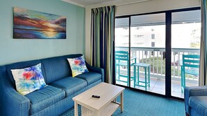 Quarto com sofá azul, uma pequena mesa de madeira e varanda com cadeiras.