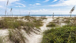Ein sandiger Pfad führt zu einem Strand mit Dünen und spärlicher Vegetation.